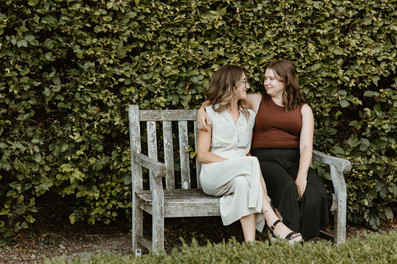 Two woman cuddle on a bench and look at each other in front of a large hedge