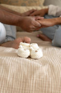 A pari of baby booties rest on a bed and in the background are the holding hands of a couple sitting crossed legged on the bed