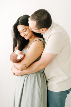 A woman stands a cradles a newborn baby in her arms while a man stands behind her and hugs her and kisses her cheek