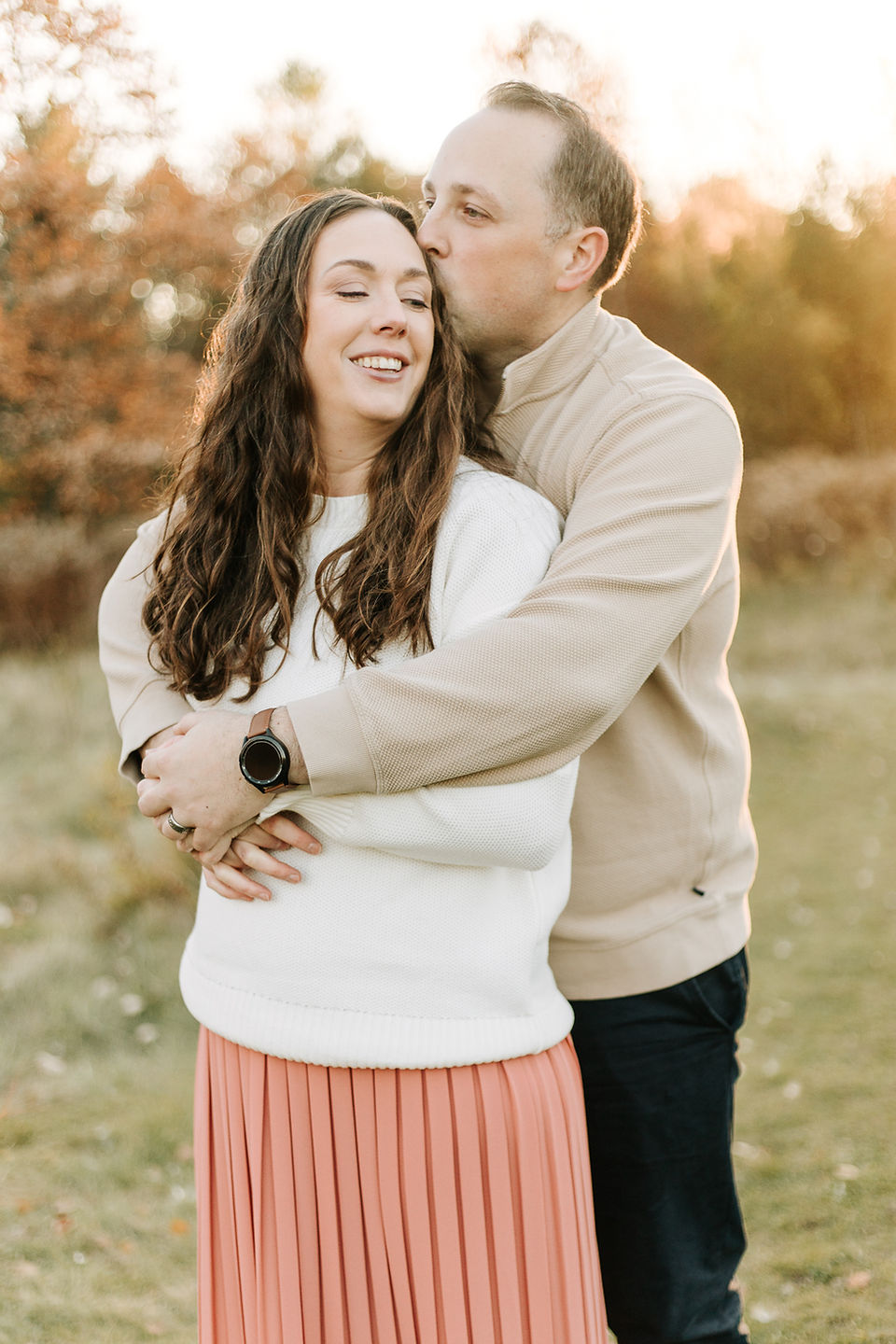 A man hugs a woman from behind and kisses her head as they smile and stand on a grass path by trees as the sun sets