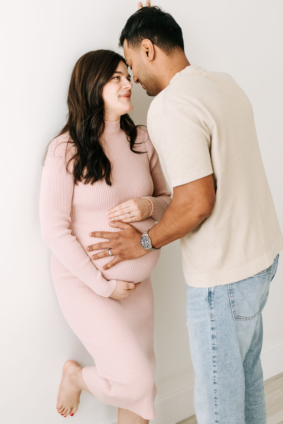 woman in a pink dress cradles pregnant stomach and leans against a wall with one foot up while looking up at a man who leans into her and puts hand on her stomach