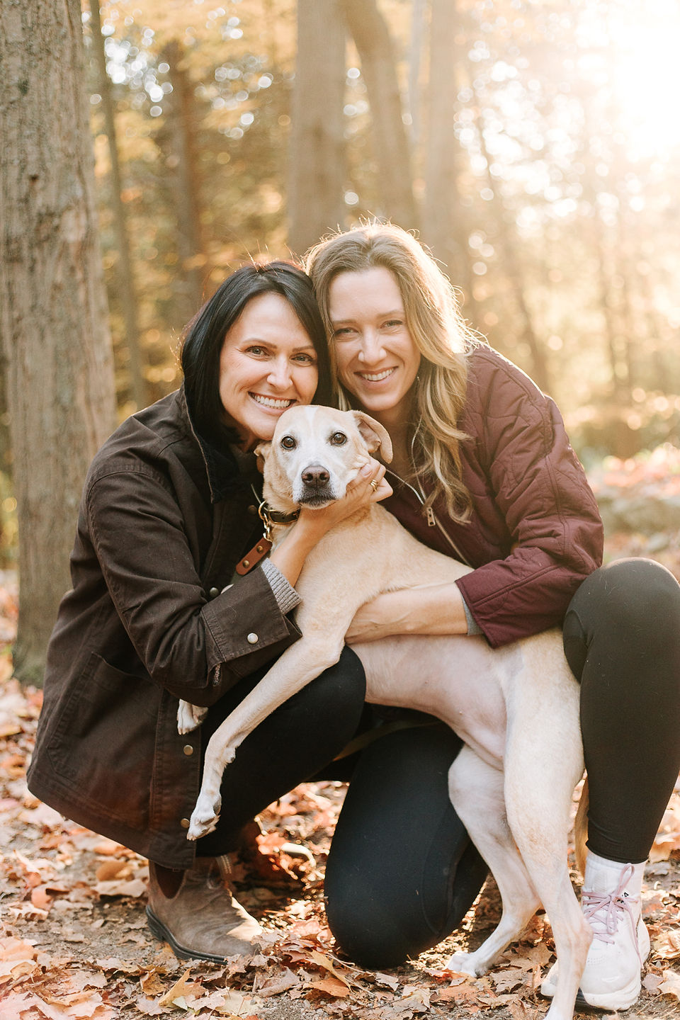 Two woman hug a dog in a forest during golden hour