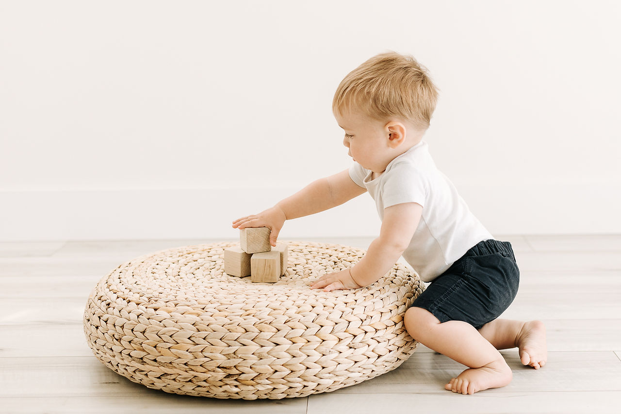 A one year old boy kneels next to a rattan pouf and plays with small wooden blocks on it
