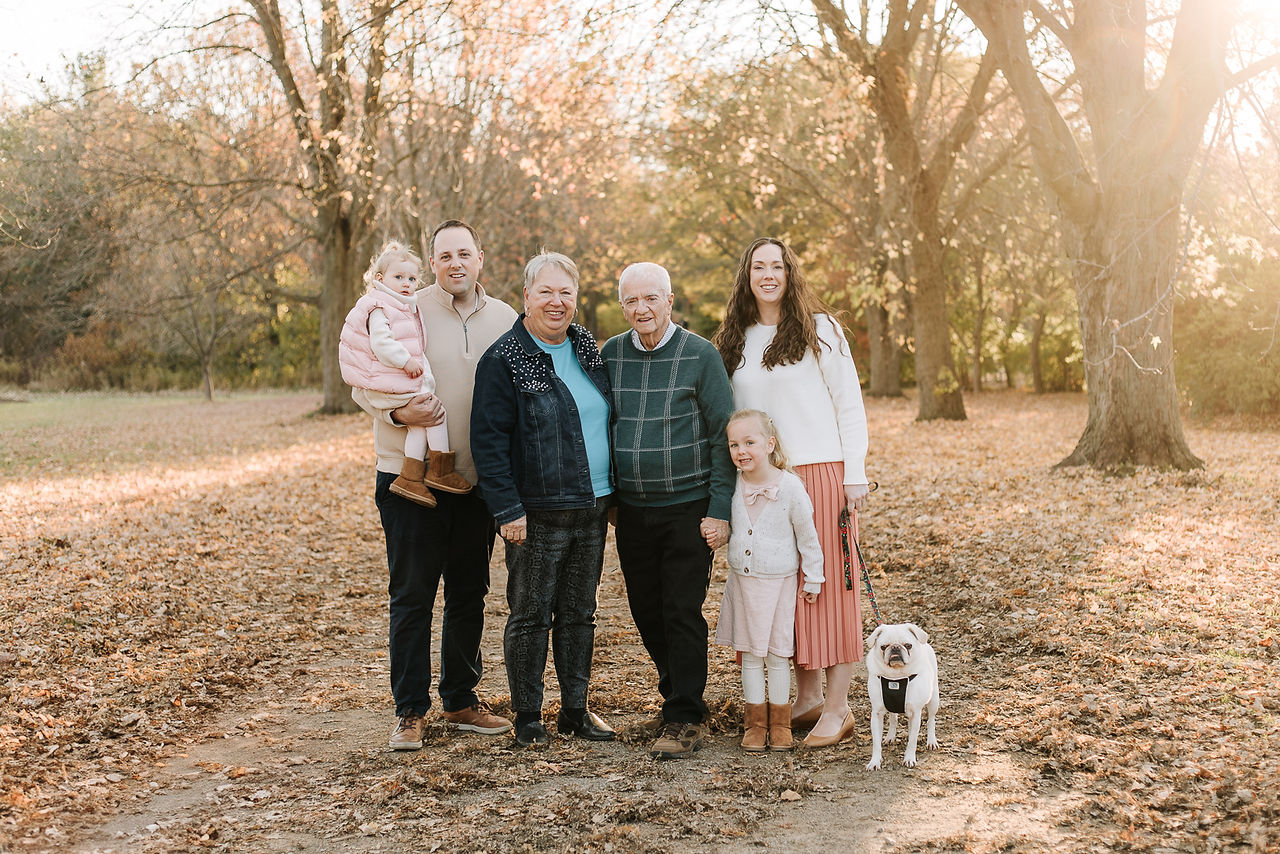 A couple stand and smile in a forest with two girl, a dog, and an older couple
