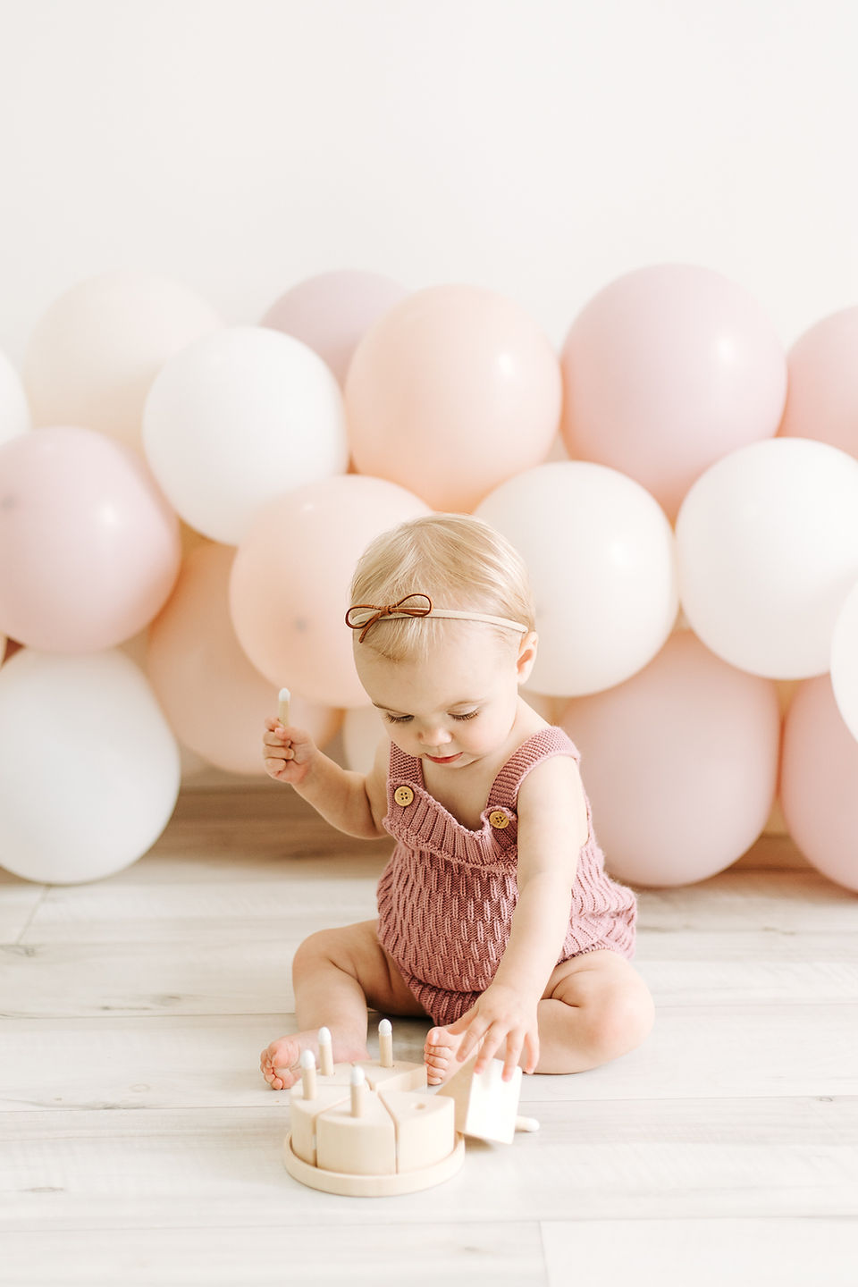 A one year old sits on the floor in front of a balloon arch and plays with a wooden cake toy