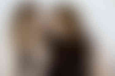 Two women sit together as one rests her back against the other as they look at each other and smile