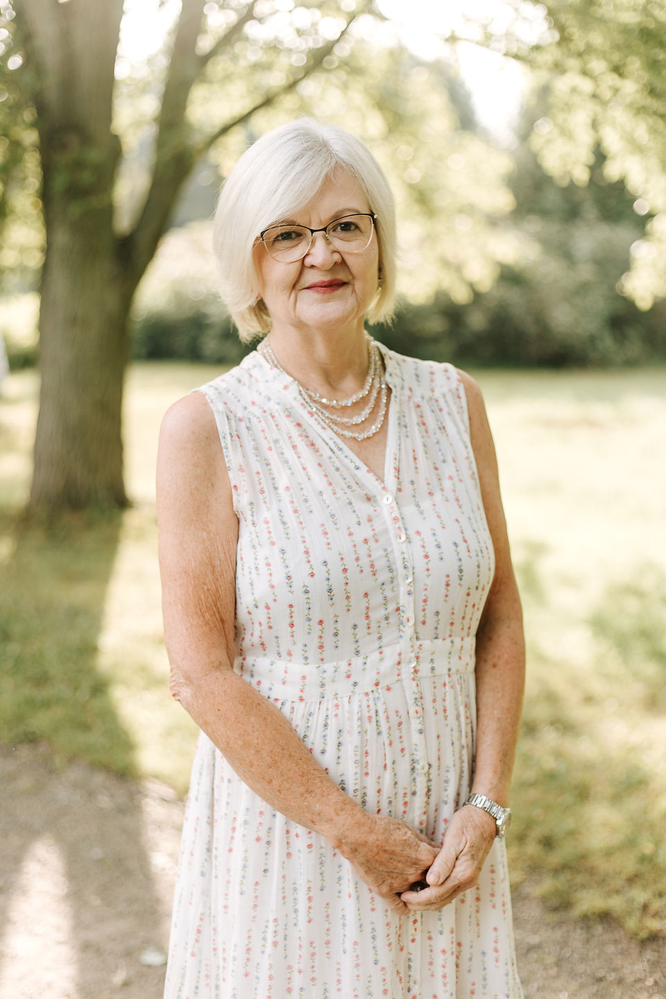 An older woman in a white dress stands on a path under trees