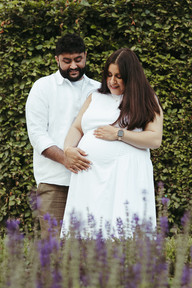 A man stands behind a pregnant woman as they both rest their hands and looks down at her stomach and stand behind lavendar