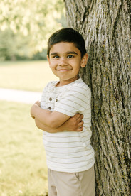 A young boy leans against a tree trunk and smiles with his arms folded