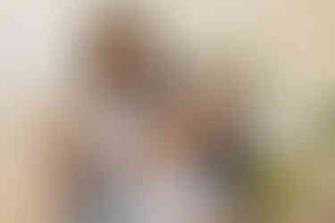 A woman in jeans and white t shirt sits on the floor laughing as a girl in white t shirt and jeans stands on her lap and sticks out her tongue