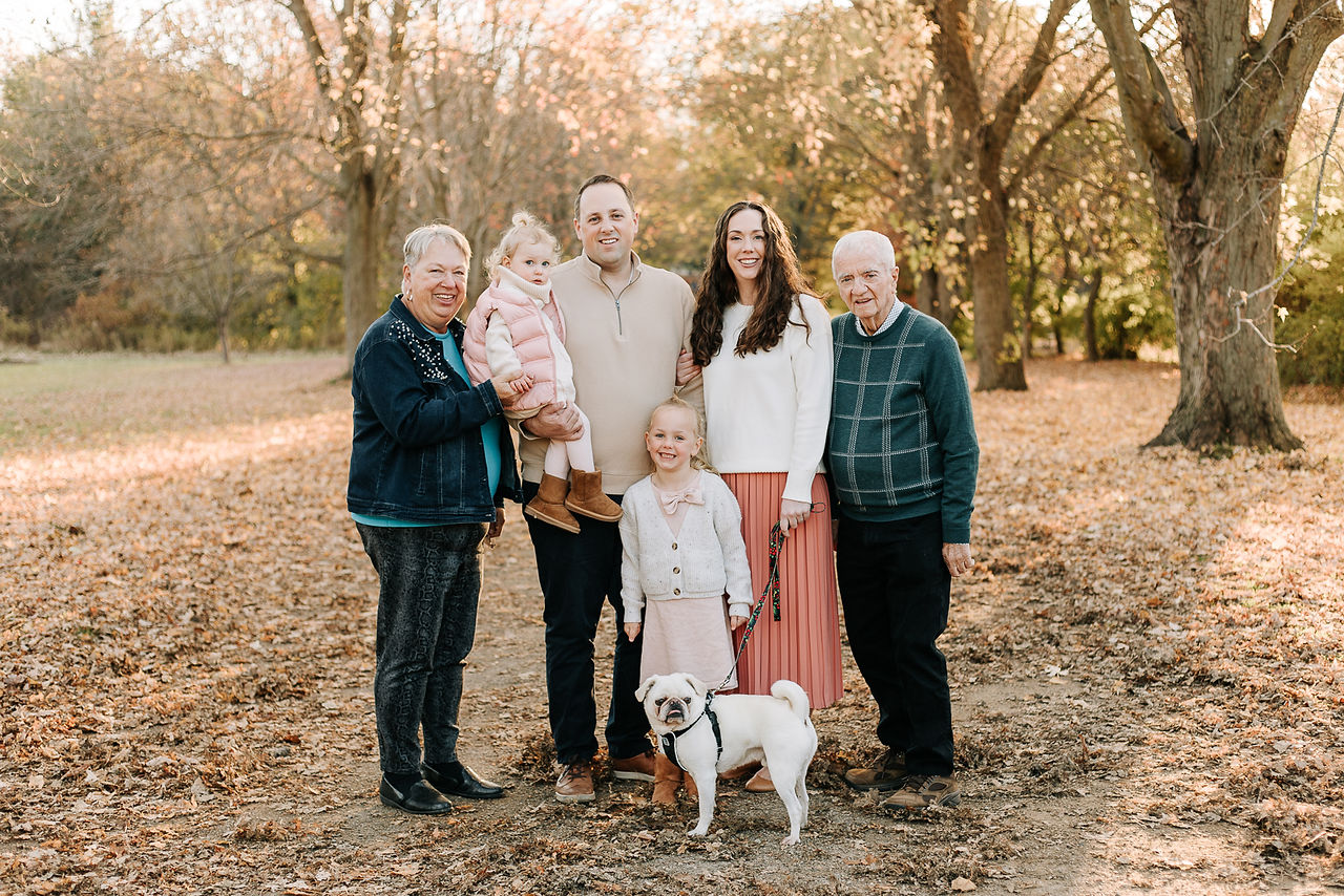 A couple stand and smile in a forest with two girl, a dog, and an older couple