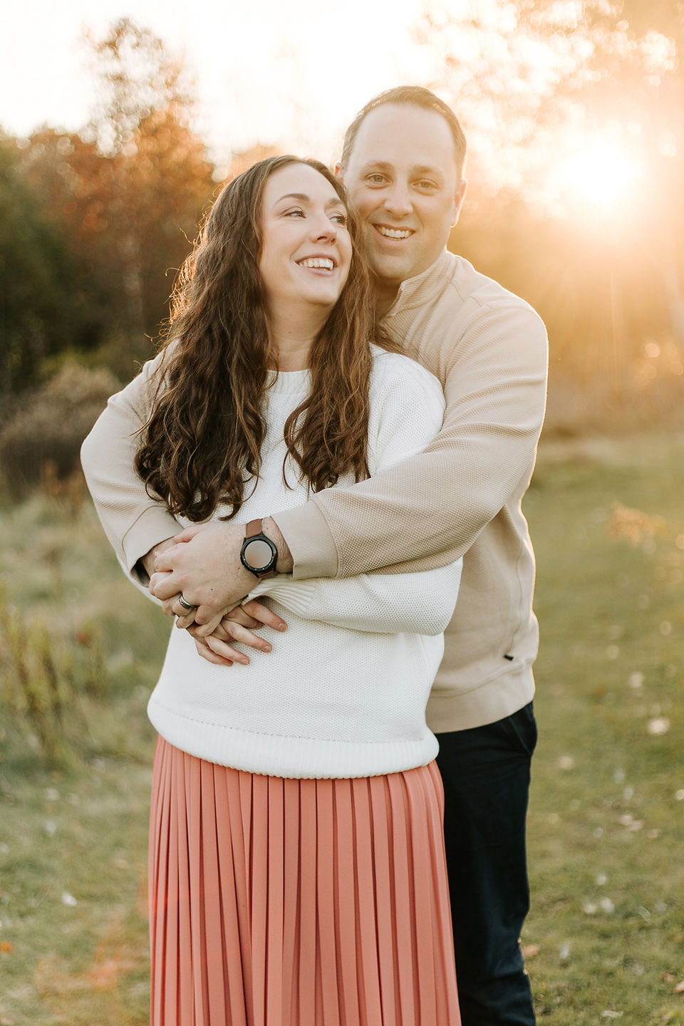 A man hugs a woman from behind as they smile and stand on a grass path by trees as the sun sets