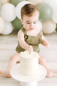 A one year old boy sits on the floor and touches a little white cake as green and white balloon arch is behind him
