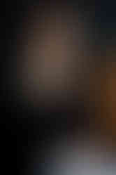 A woman in a black top and black jacket sits on a brown couch and smiles