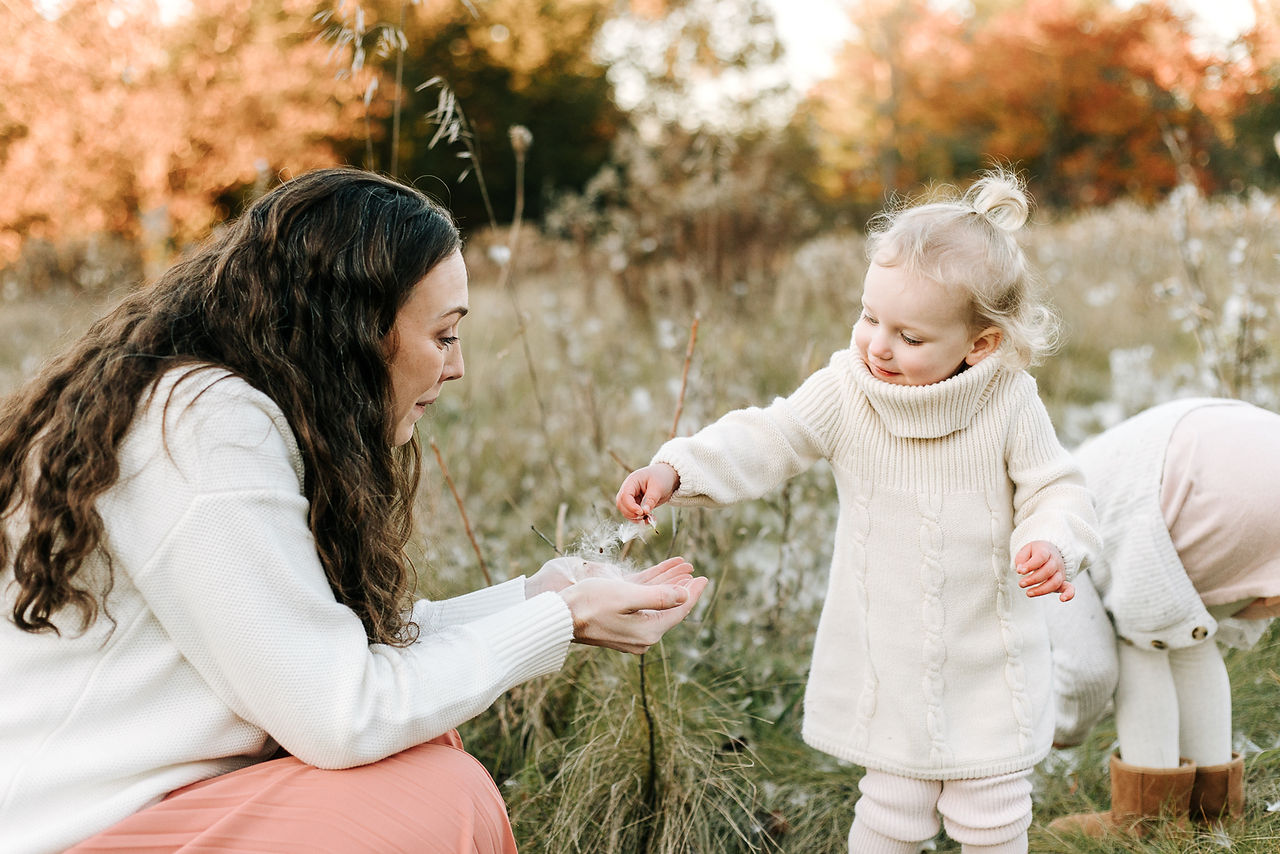 A woman crouches down and smiles at a young girl as they stand in a grass field with milkweed