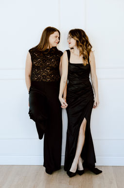 Two woman hold hands and look at each other as they lean against a white wall