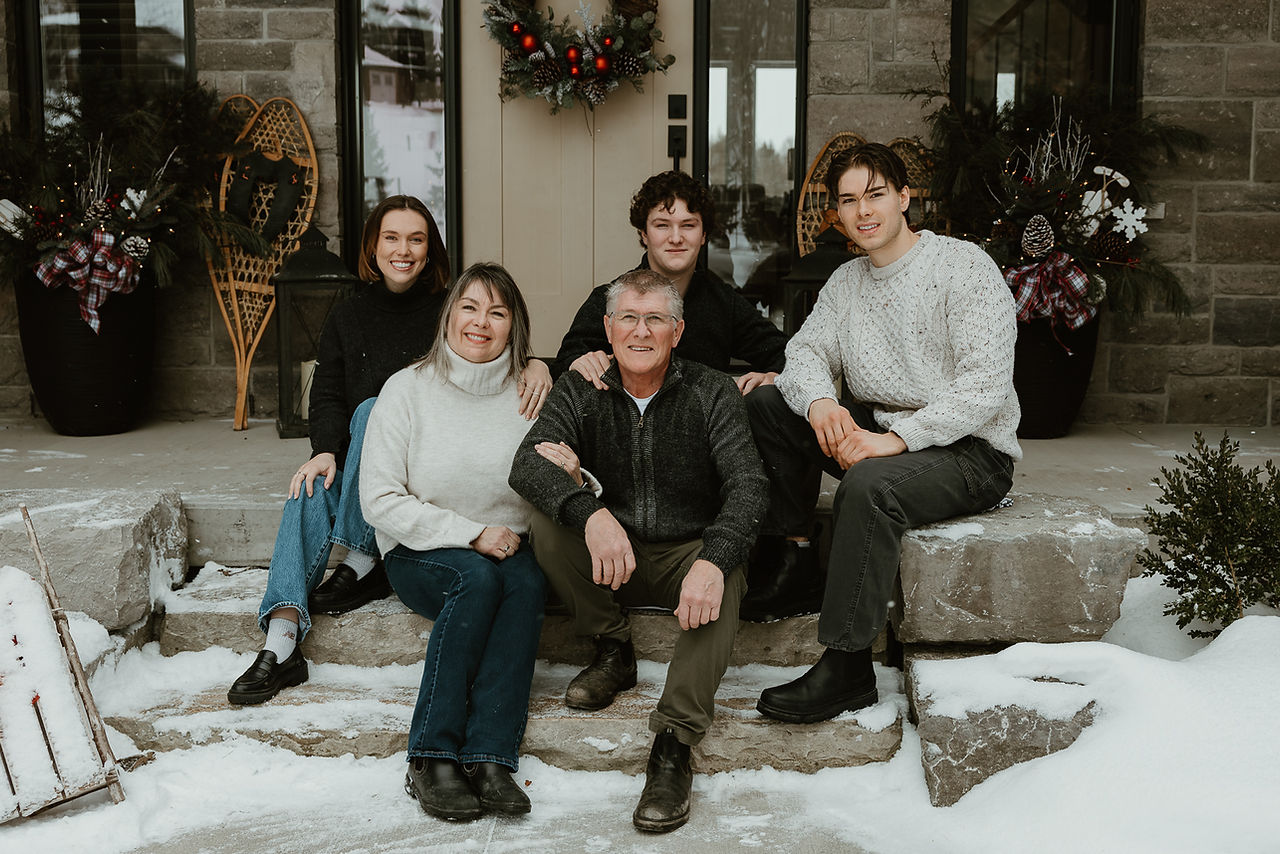 A family of five with grown children sit on a doorstep in winter and smile