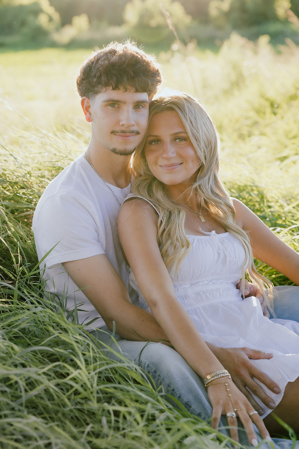 A teen boy and girl sit together on grassy field and rest their heads together