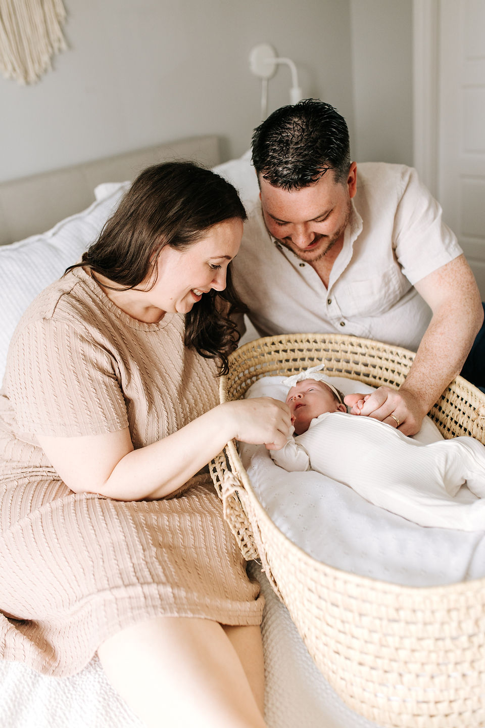 A woman and a man sit on a bed with a newborn baby in a moses basket laying between them