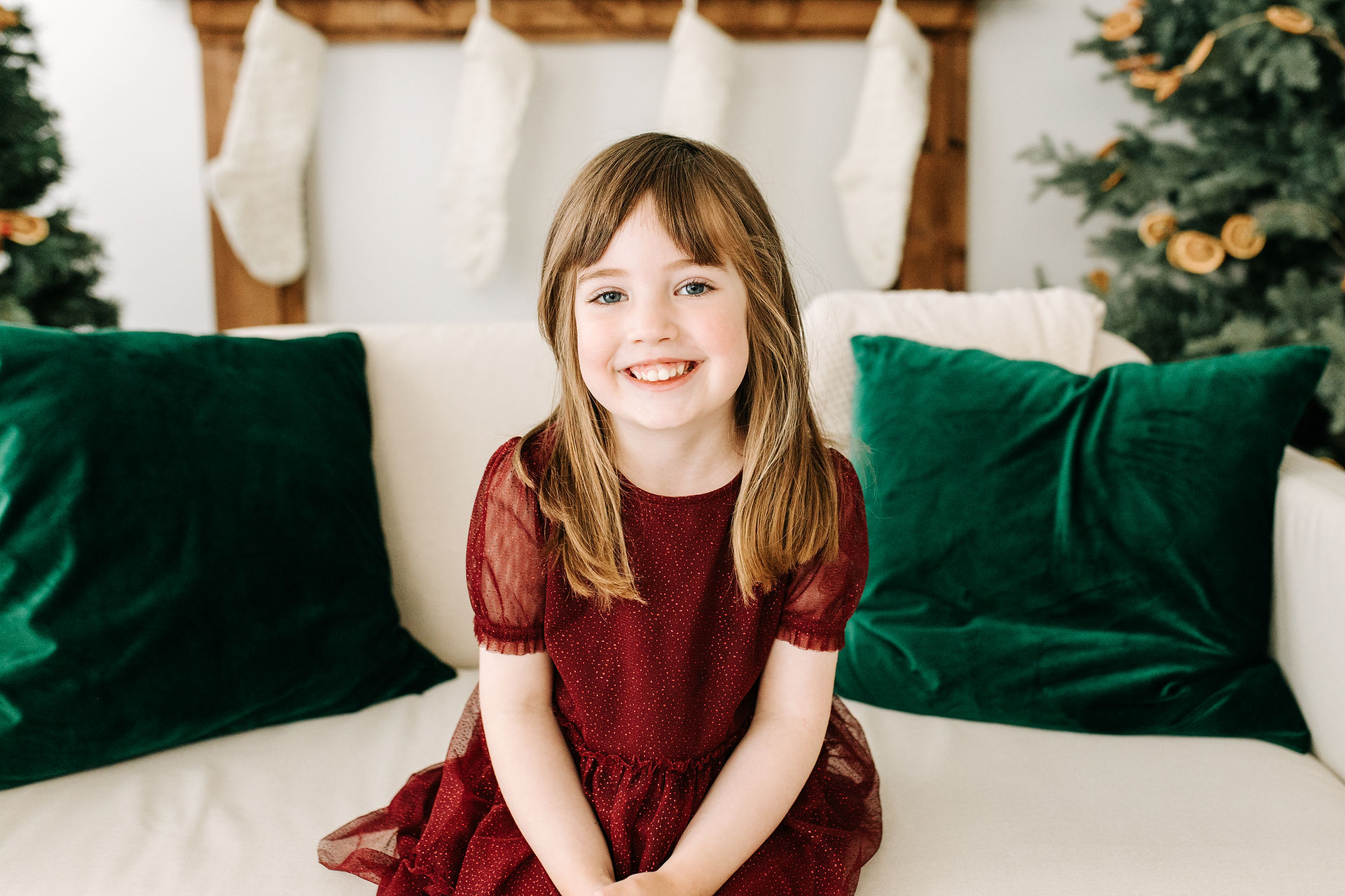 A young girl in a red dress sits on a couch in between two Christmas trees