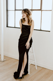 A woman in a long black dress sits on a stool and looks into the distance