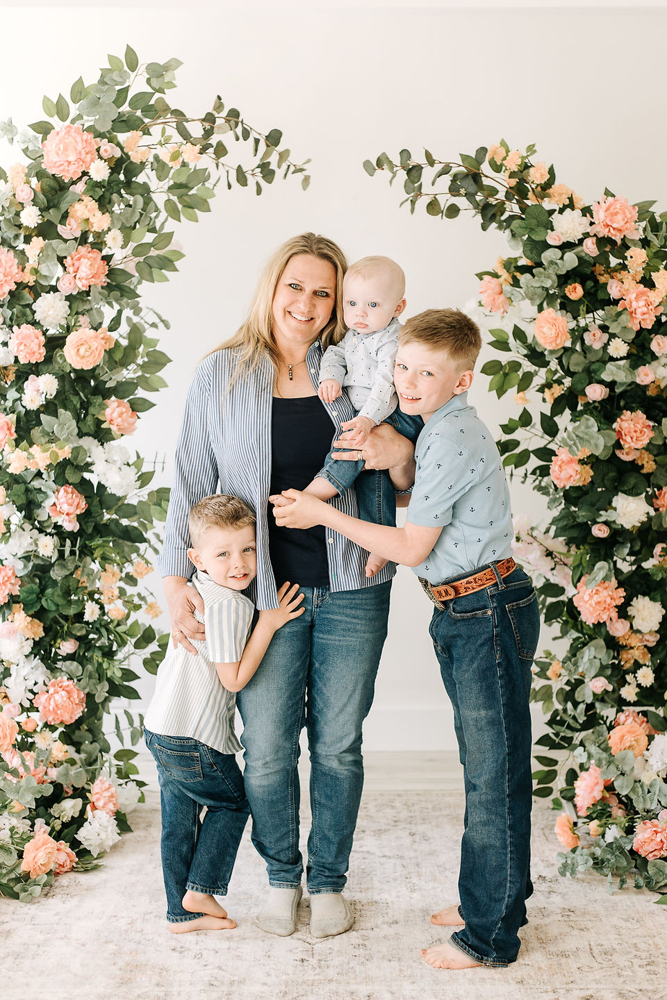 A woman holds a baby in her arms with a boy standing on either side of her hugging her with flowers behind them
