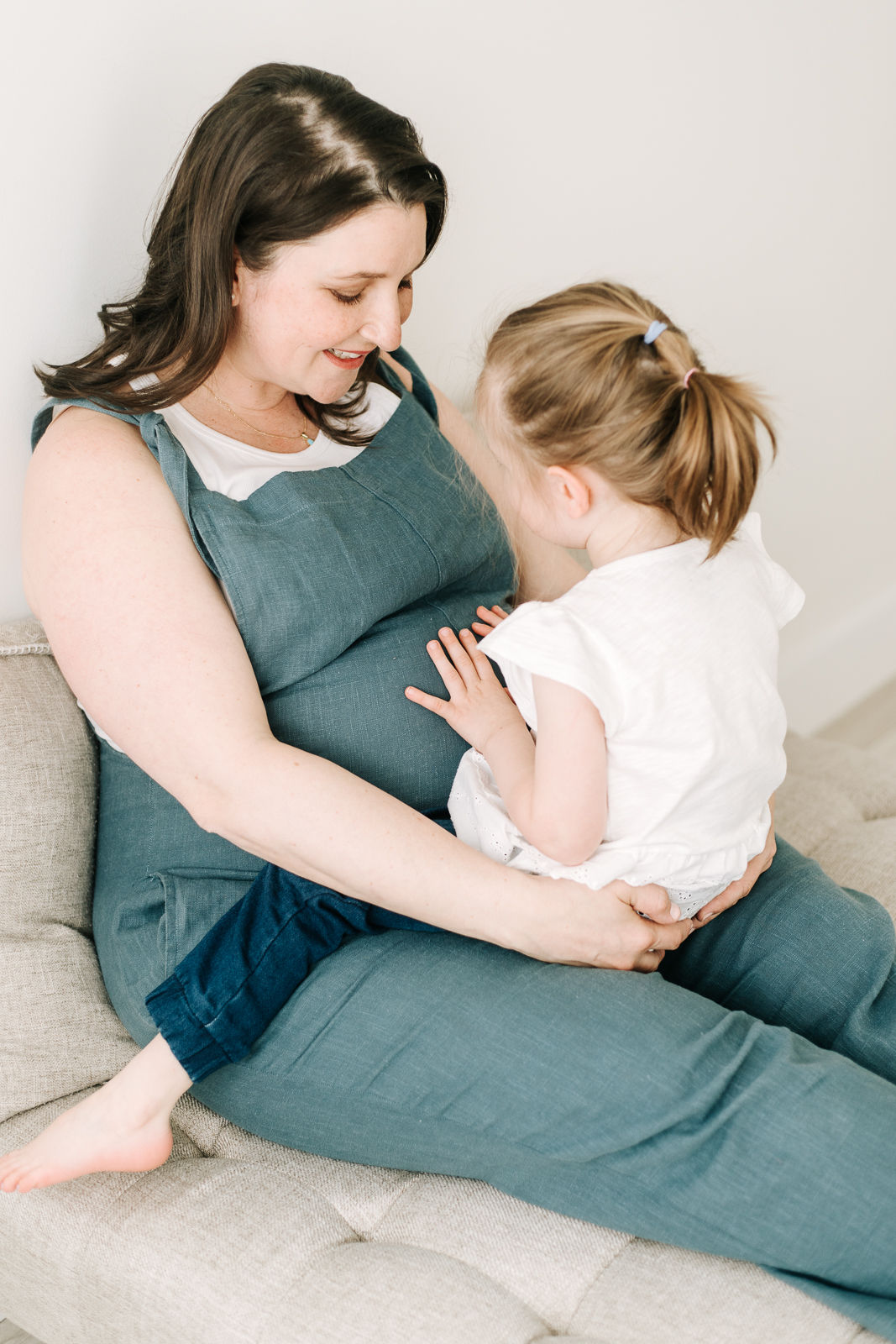 A pregnant woman in blue overalls sits on a couch with a young girl on her lap who is resting her hands on the woman's stomach