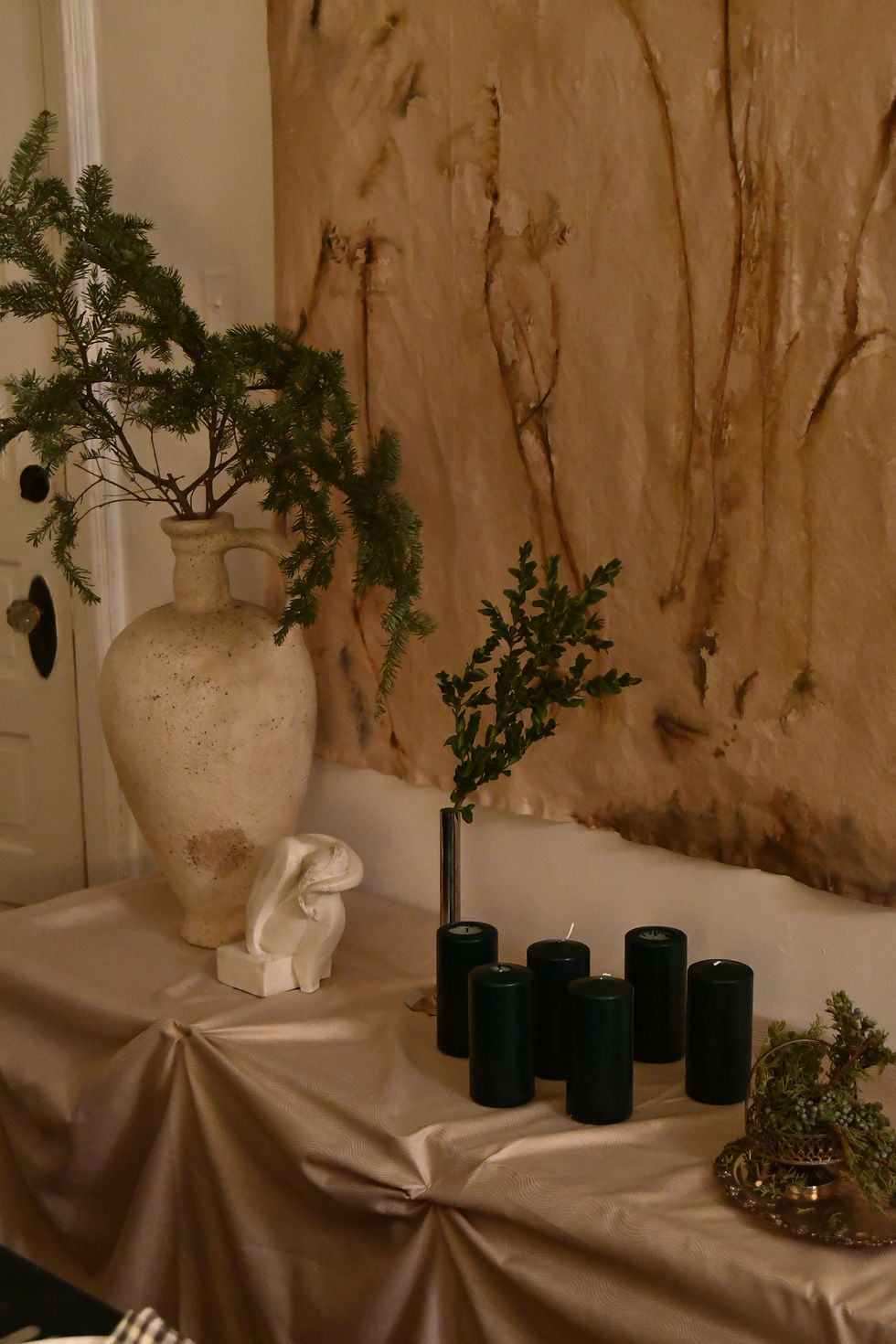 A serene setting with a large beige vase holding greenery, black candles on a table with a draped cloth, and a textured wall backdrop.