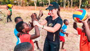 Woman in black cap high-fives smiling children on a dry outdoor field; one boy holds a colorful ball, playful and cheerful.