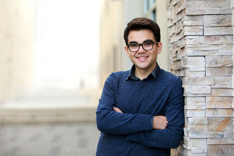 Headshot of a young man wearing glasses and a blue sweater leans against a stone wall