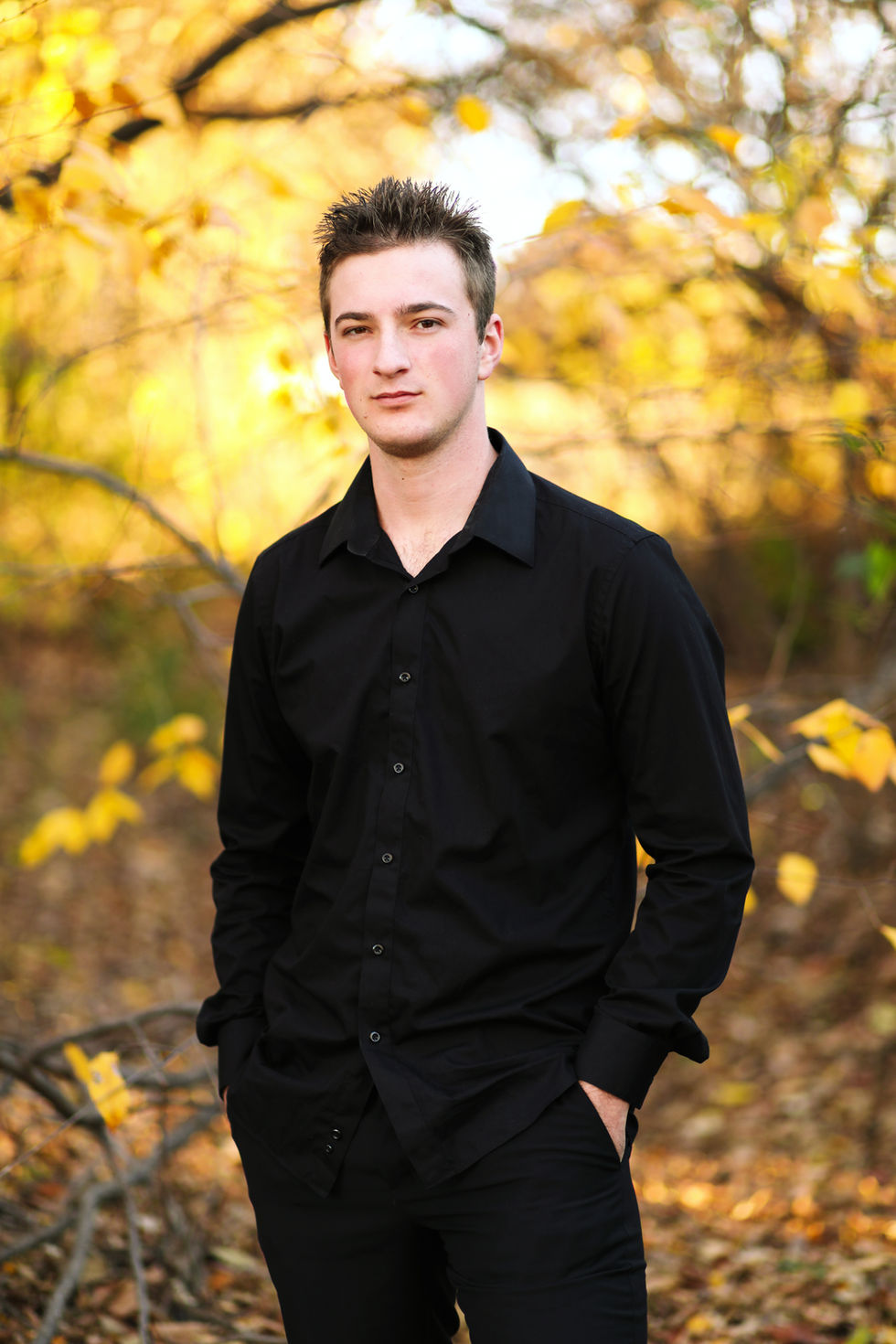 portrait of a young man in a black shirt stands with his hands in his pockets
