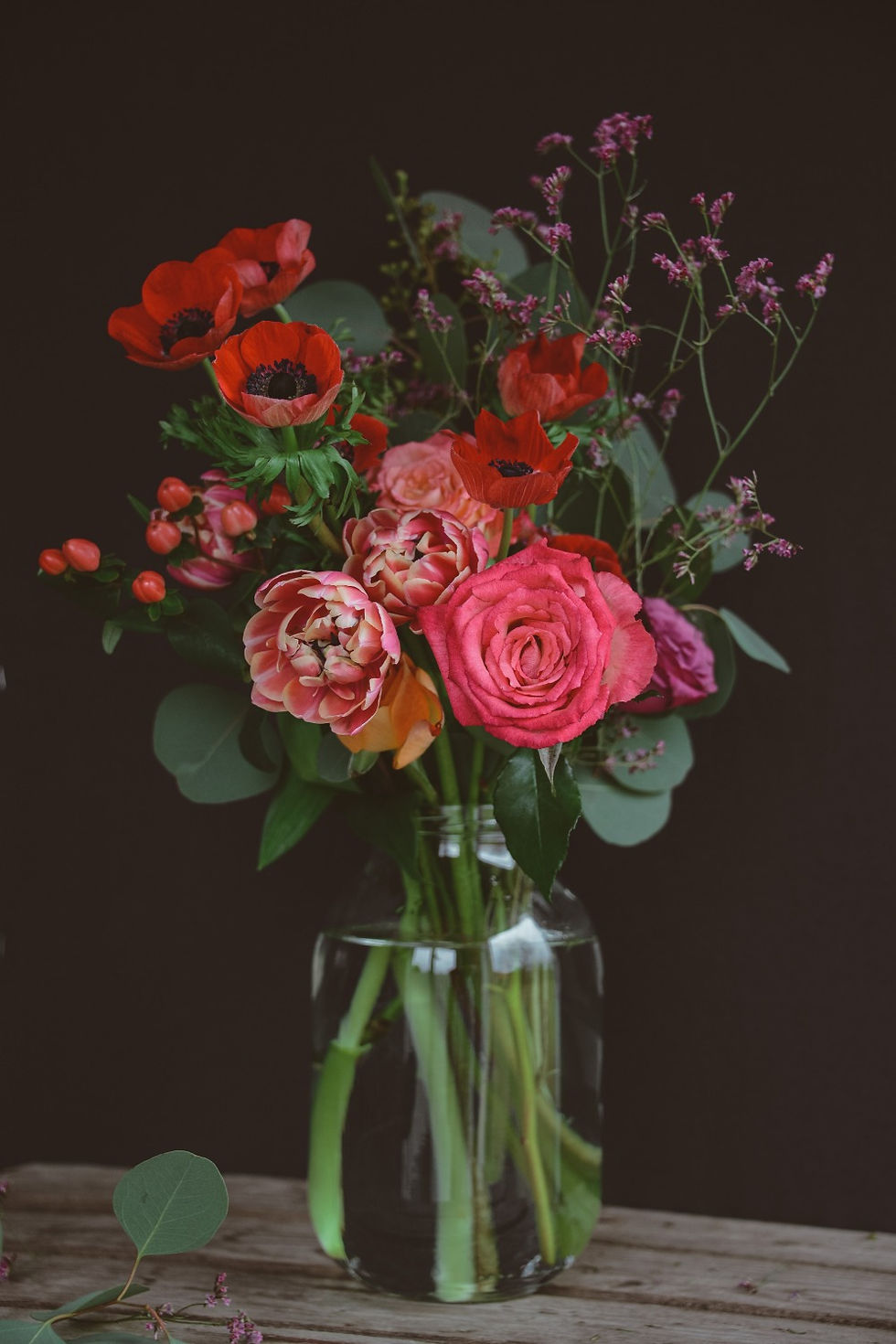 An almost-black background. On a table, a clear glass vase with a bouquet of red and purple flowers, and eucalyptus leaves.