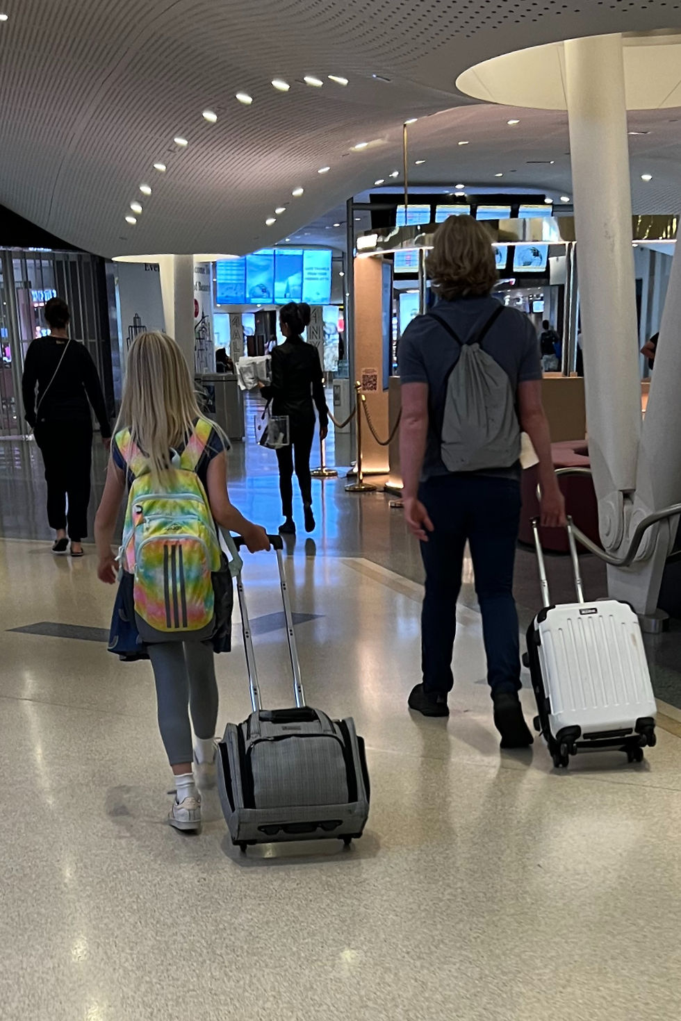 girl and a young man walk away through an airport terminal, each wheeling a roller suitcase past duty-free shops