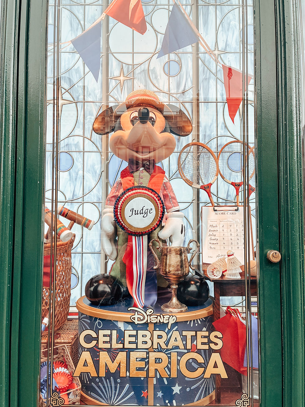 Mickey Mouse dressed as a Judge in a patriotic red, white, and blue outfit with straw boater hat, standing in a festive display at the Emporium on Main Street USA in Magic Kingdom. He is surrounded by croquet mallets, badminton rackets, and trophies as part of Disney's celebration of America's 250th birthday.