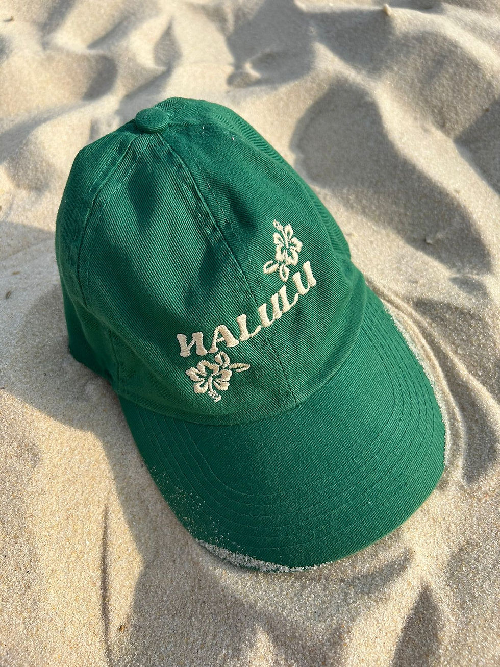 Boné verde com bordado floral e a palavra 'HALULU' na frente, repousando na areia da praia