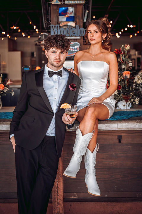 Bride and groom at outdoor, covered bar. Bride sitting up on the bar, groom holding a drink.