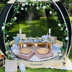 Picnic setup with tent, flowers, white roses, and text #Kiyosfunfair on the green lawn.