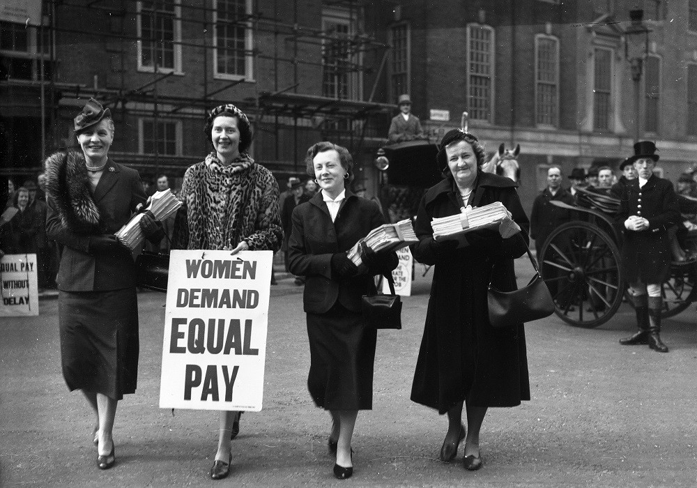 Black-and-white photo of four well-dressed women campaigners. One is carrying a placard with the text 'Women Demand Equal Pay'.