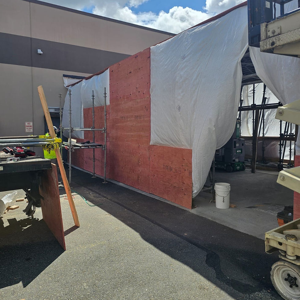 Construction site with red plywood wall, white tarp, scaffolding, equipment carts, bucket, and a No Parking sign.