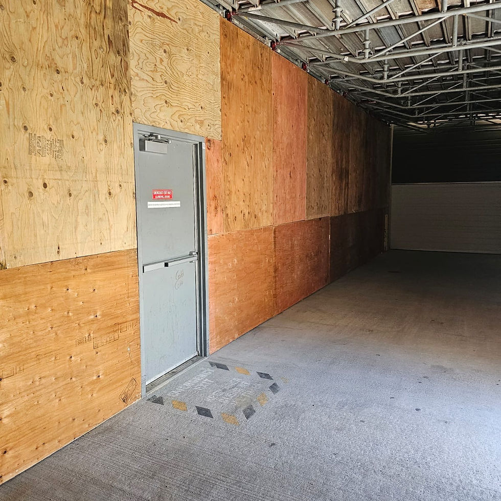 Boarded-up industrial corridor with a gray emergency exit door, plywood walls, and exposed metal ceiling.