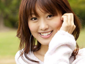 Asian woman with a white sweater on smiling at the camera with a white string coming from her ears. She has brown hair with bangs. She is outside and the background is blurry.