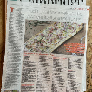 A newspaper titled "Cambridge" features an article on traditional flammekueche with a recipe and colorful dish photo. Rustic wooden table setting.