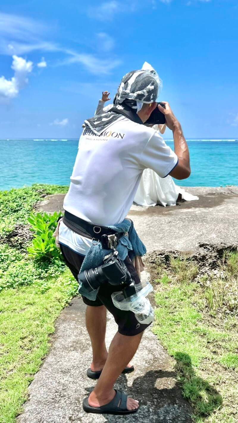 沖縄の青い海を背景に、フォトウエディングのカメラを構えるカメラマンのYashiki。撮影の瞬間を捉えた和やかな雰囲気。