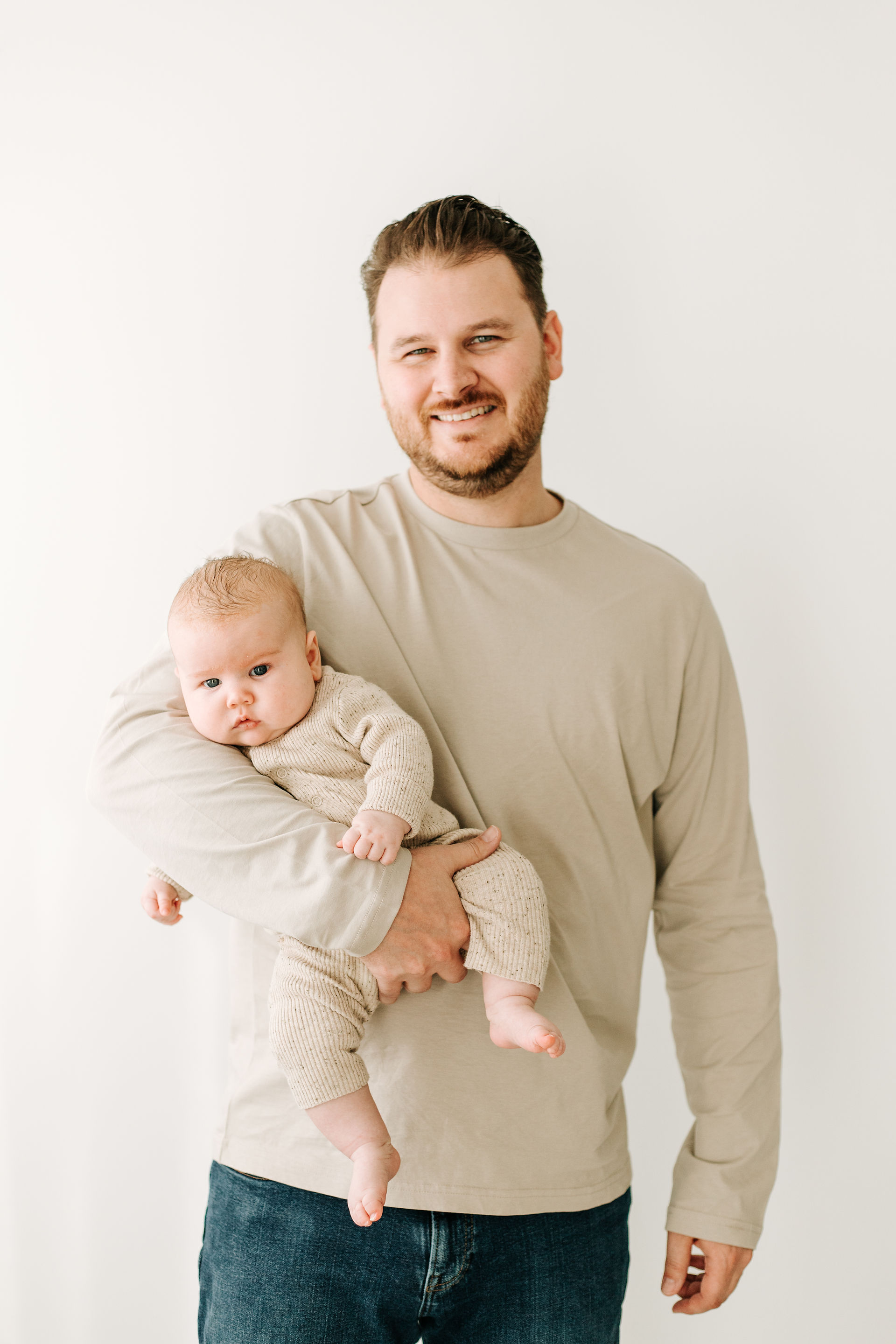A smiling father in a beige long-sleeve shirt and jeans holds his baby in one arm. The baby, wearing a matching knit outfit, looks alert and cozy. The portrait is taken against a clean, light background.