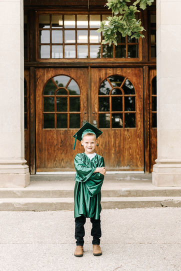 Kindergarten student standing in front of school doors wearing green graduation cap and gown