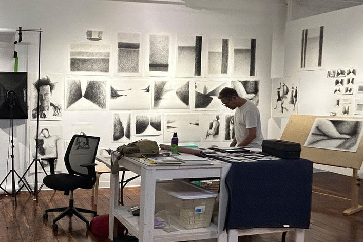 artist Douglas Gilbert standing in front of a table in his studio. Many drawings on the wall.