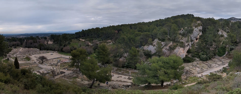 A panorama of the ruins of an ancient city. The ruins are in a small valley between tree-filled hills. The sky is mostly cloudy.