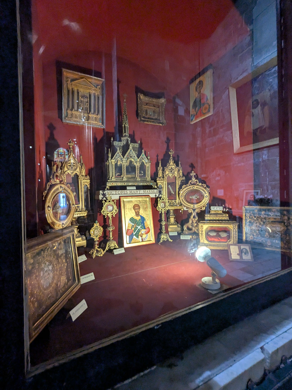 A glassed display case containing many saint reliquaries. The background is red, the reliquaries give off a mainly golden glow.