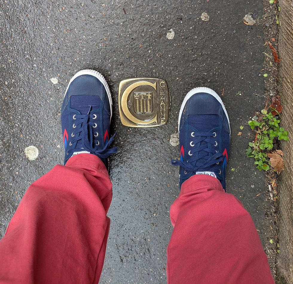 A medallion embedded in the sidewalk with a "C" and the name "Cezanne" on it. Two feet in blue sneakers attached to two legs in red pants are next to the medallion.