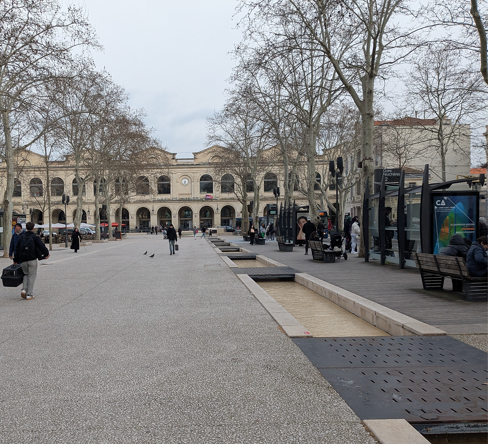 A pedestrian boulevard with a small water channel built into the concrete walkway. There is a train station building at the end of the street. A few trees line the street, and there are a few people walking.